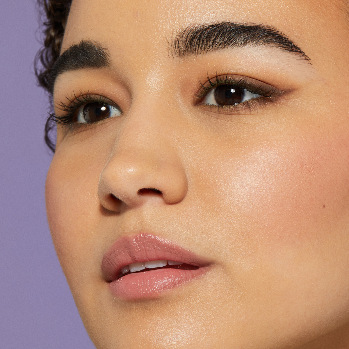 Close-up of a person with clear skin, glossy pink lips, and defined brows, against a purple background.