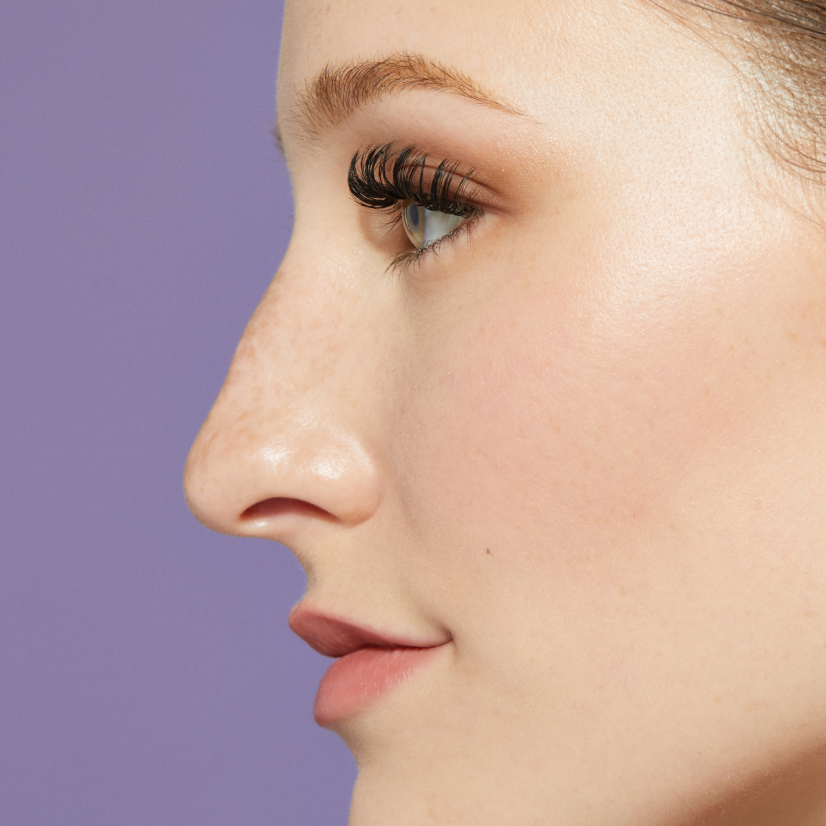 Close-up side profile of a person with fair skin, freckles, and long eyelashes against a purple background.